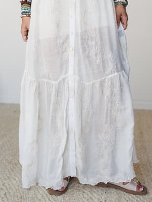 A close-up of a flowy white maxi dress covered in delicate floral embroidery, styled with layered beaded bracelets and flat sandals — the kind of dress that feels equally at home at a beach wedding or a slow summer morning.