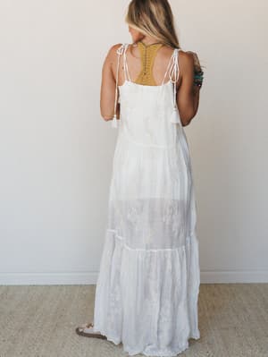A white embroidered maxi dress shot from the back, with tiered layers and tie shoulders — the kind of dress that feels equally at home at a beach wedding or wandering a coastal market.