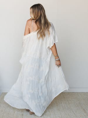 A flowing white maxi kimono with floral embroidery catches mid-twirl, shot from behind — the kind of piece that looks just as good doing nothing as it does everything.