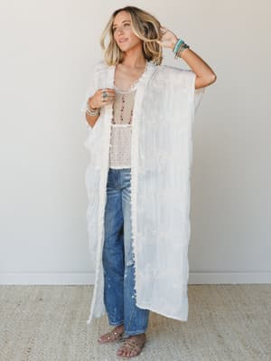A flowy, floor-length sheer kimono with floral embroidery is layered effortlessly over a beaded cami and distressed wide-leg jeans — the kind of outfit that looks like it came together on purpose without trying too hard.