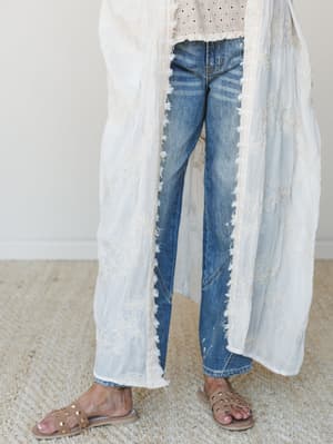 A relaxed, romantic layering look — an embroidered cream kimono draped open over a dotted top and distressed straight-leg jeans, finished with studded tan slide sandals on a neutral carpet backdrop.