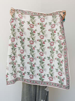 A model holds up a large block-print floral scarf or bandana in ivory with trailing botanical vines in crimson and sage, bordered by an intricate printed edge — the kind of piece that does three things at once and looks effortless doing all of them.