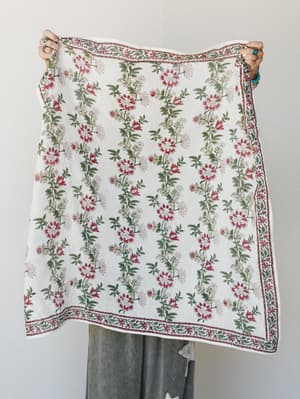 A model holds up a large block-print floral scarf or bandana in ivory with trailing botanical vines in crimson and sage, bordered by an intricate printed edge — the kind of piece that does three things at once and looks effortless doing all of them.