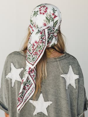 Shot from behind, a model wears a floral block-print bandana tied over her head, trailing down her back alongside loose blonde hair, paired with an oversized washed grey tee featuring distressed white star appliqués — the kind of outfit that looks ef