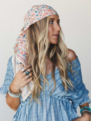 A sun-warmed studio shot of a model wearing a floral printed bandana tied loosely over her head, paired with a chambray off-shoulder top and layered turquoise and silver jewelry — the whole look has that effortless, road-trip-ready energy.