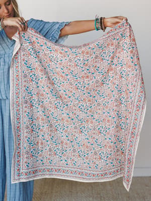 A model holds up a large square block-print bandana with an all-over floral vine pattern and decorative border, styled against a neutral interior backdrop — the kind of piece that looks equally at home as a scarf, wrap, or wall hanging.