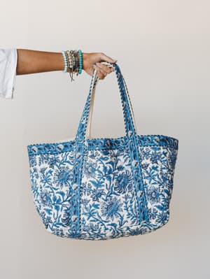 A hand holds up a large quilted block-print tote in ivory and cobalt blue floral — stacked beaded bracelets on the wrist, the whole thing feeling like it already has a farmers market run and a beach day planned.