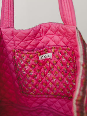 A close-up of a quilted tote bag in hot pink with a small floral block-print front pocket, showing diamond stitching detail and a woven 'POL' label — the kind of bag that makes you want to pack it immediately.