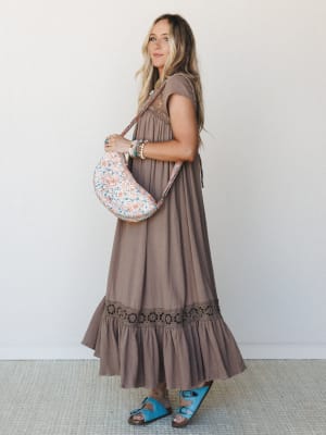 A flowy mocha maxi dress with crochet trim and a ruffled hem is styled with a floral crossbody bag and turquoise sandals — the kind of outfit that looks effortless but was absolutely thought about.