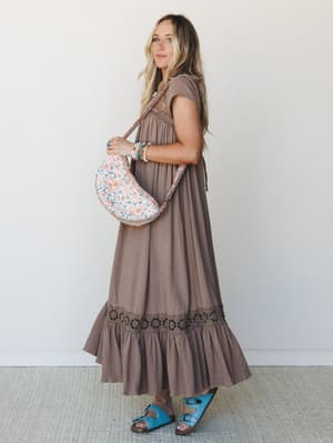 A flowy mocha maxi dress with crochet trim and a ruffled hem is styled with a floral crossbody bag and turquoise sandals — the kind of outfit that looks effortless but was absolutely thought about.