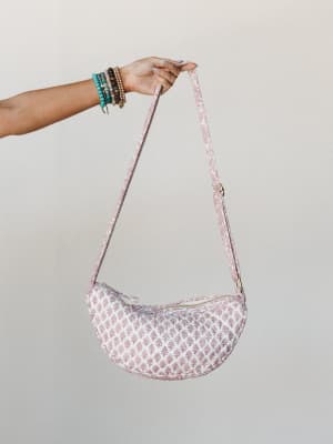A crescent-shaped quilted crossbody bag in cream and dusty rose block print is held aloft by a stacked-bracelet wrist, shot clean against a neutral studio wall — the kind of bag that makes a casual outfit feel like a decision.