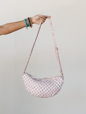 A crescent-shaped quilted crossbody bag in cream and dusty rose block print is held aloft by a stacked-bracelet wrist, shot clean against a neutral studio wall — the kind of bag that makes a casual outfit feel like a decision.
