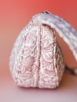 A close-up side profile of a quilted fabric bag in a soft ivory and dusty rose floral print, shot against a warm coral-to-blush gradient background — the kind of bag that makes a whole outfit feel more intentional.