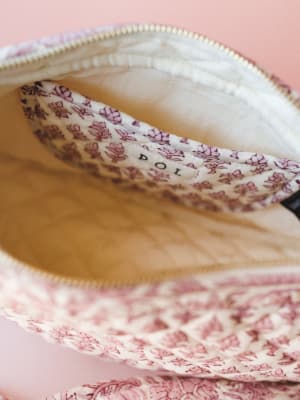 A close-up detail shot of an open quilted pouch in ivory and dusty rose, revealing a sweet floral-printed interior pocket with a brand label — the kind of bag that makes you happy every time you unzip it.