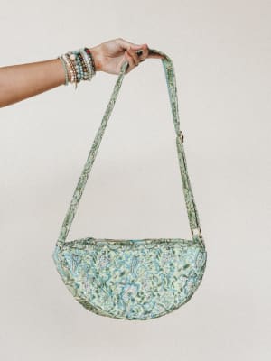 A hand adorned with stacked beaded bracelets holds up a crescent-shaped quilted crossbody bag in a soft pistachio green with blue and white floral paisley print — the kind of bag that makes an outfit feel intentional without trying too hard.