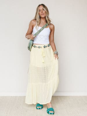 A sun-warmed outfit with easy summer energy — white eyelet crop tank, flowy pale yellow maxi skirt with fish embroidery and tassel tie waist, styled with a green quilted crossbody bag and teal beaded sandals.