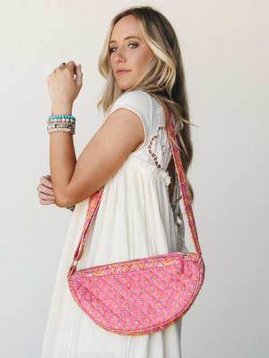 A model posed three-quarter back, showing off a half-moon quilted crossbody bag in bright pink with a small floral block print — worn over a flowy white dress with crochet back detail and stacked beaded bracelets that feel like a summer afternoon wit