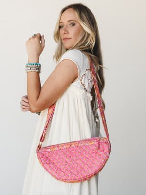 A model posed three-quarter back, showing off a half-moon quilted crossbody bag in bright pink with a small floral block print — worn over a flowy white dress with crochet back detail and stacked beaded bracelets that feel like a summer afternoon wit