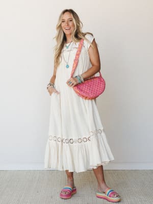 A breezy cream midi dress with floral crochet trim and a ruffled hem is styled with a bright pink quilted crossbody bag, rainbow platform sandals, and turquoise jewelry — the kind of outfit that feels equally at home at a farmers market or a beach to