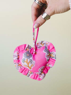 A hand adorned with silver rings holds up a fabric heart charm in bright fuchsia and floral print, ruffled at the edges and finished with a looped hanging strap — sweet, handmade-feeling, and very much the kind of thing you'd emotionally attach to a