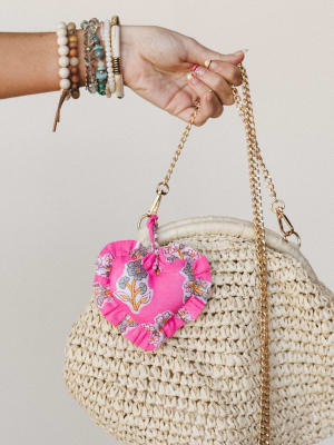 A stacked-bracelet wrist holds up a woven straw tote by its gold chain strap, with a ruffled heart-shaped bag charm in hot pink floral stealing the whole show.