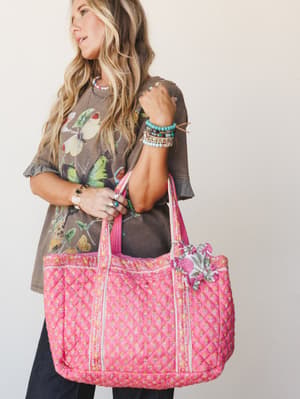 A sun-kissed model carries an oversized quilted pink tote layered with a butterfly graphic tee, stacked turquoise bracelets, and rings — the whole look has that effortless, collected energy of someone who always has the best bag at the farmers market