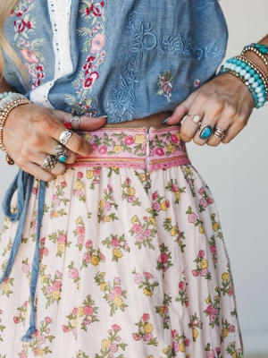 A close-up waist shot capturing the layered detail of a flowy floral skirt with a printed ribbon waistband paired with an embroidered denim crop top, styled with stacked turquoise and silver jewelry — the kind of outfit that looks like it was put tog