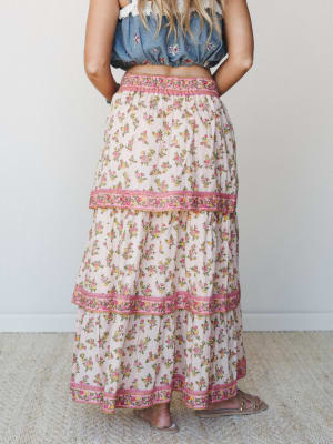 A flowy tiered maxi skirt in a ditsy floral print — cream base with pink and yellow blooms — styled from the back with a denim crop top and flat sandals, giving off that effortless warm-weather energy.