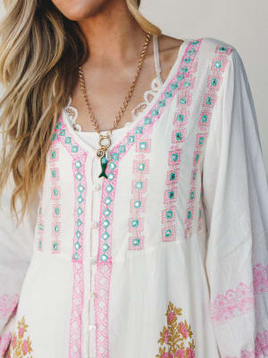 A close-up of a white boho blouse or dress layered over a white lace camisole, covered in pink geometric embroidery and turquoise mirror embellishments — the kind of piece that looks like it came back from somewhere more interesting than you have.
