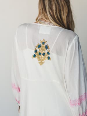 A back-facing close-up of a flowing ivory maxi dress, highlighting a hand-embroidered floral motif in gold and teal at the center back, with pink block-print trim along the long sleeves — the kind of piece that looks like it came home from somewhere