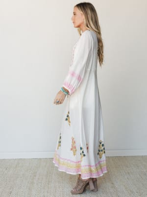 A flowy white maxi dress with colorful block-print floral motifs along the skirt and cuffs — the kind of dress that makes you want to book a flight somewhere warm.