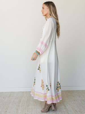 A flowy white maxi dress with colorful block-print floral motifs along the skirt and cuffs — the kind of dress that makes you want to book a flight somewhere warm.