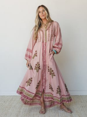 A flowy block-print maxi dress in dusty pink with floral embroidery, mirror embellishments, and a border hem — styled with layered necklaces, turquoise bracelets, and flat sandals for a well-traveled, effortlessly personal look.