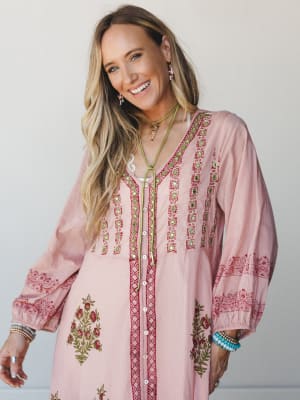 A smiling model in a blush pink maxi dress covered in intricate block-print florals, mirror embellishments, and crimson embroidery — styled with layered bracelets and a green cord necklace for that perfectly collected, free-spirited feel.