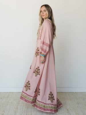 A flowy pink maxi dress with intricate floral block-print embroidery along the hem and sleeves, styled with turquoise stacked bracelets and a turquoise ring — the kind of dress that makes a Tuesday feel like a little trip somewhere warm.