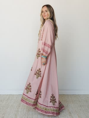 A flowy pink maxi dress with intricate floral block-print embroidery along the hem and sleeves, styled with turquoise stacked bracelets and a turquoise ring — the kind of dress that makes a Tuesday feel like a little trip somewhere warm.