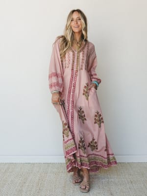 A flowy block-print maxi dress in dusty rose with olive and cranberry floral motifs, styled with stacked bracelets and strappy sandals against a clean studio wall — effortless and well-traveled.