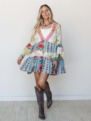 A flowy mixed-print mini dress in cream and dusty blue with bold floral panels, styled with dark brown cowboy boots and stacked bracelets — free-spirited and a little bit country, in the best way.