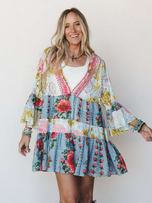 A smiling model wears a flowy, tiered mini dress in a vibrant patchwork floral print with bell sleeves and red crochet trim at the neckline, layered over a white tee and styled with stacked boho bracelets — the kind of outfit that looks like it was m