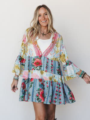 A smiling model wears a flowy, tiered mini dress in a vibrant patchwork floral print with bell sleeves and red crochet trim at the neckline, layered over a white tee and styled with stacked boho bracelets — the kind of outfit that looks like it was m