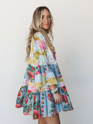A tiered mini dress in a bold patchwork floral print — light blue, pink, and golden yellow — worn by a model shot from the side against a soft white wall, with stacked bracelets and rings adding to the free-spirited, collected feel.