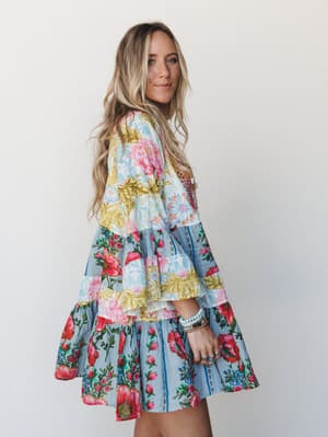 A tiered mini dress in a bold patchwork floral print — light blue, pink, and golden yellow — worn by a model shot from the side against a soft white wall, with stacked bracelets and rings adding to the free-spirited, collected feel.
