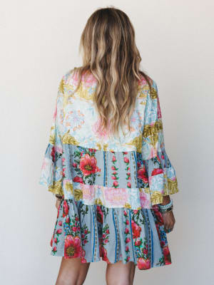 A flowy patchwork mini dress shot from behind, layered with mismatched floral prints in soft blue, cream, and bright pops of red and pink — the kind of dress that looks like a garden party decided to become a garment.