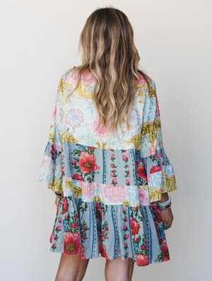 A flowy patchwork mini dress shot from behind, layered with mismatched floral prints in soft blue, cream, and bright pops of red and pink — the kind of dress that looks like a garden party decided to become a garment.