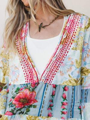 A close-up detail shot of a vibrant mixed floral dress with cowrie shell trim and red embroidered stitching along the v-neckline, layered over a white tank — the kind of piece that looks like it was found on a good trip and never given back.