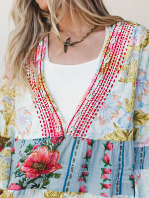 A close-up detail shot of a vibrant mixed floral dress with cowrie shell trim and red embroidered stitching along the v-neckline, layered over a white tank — the kind of piece that looks like it was found on a good trip and never given back.
