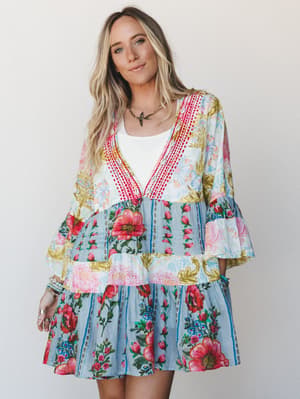 A flowy tiered mini dress in a vibrant mixed floral print — the kind of piece that looks like it was made for wandering a farmers market or dancing at an outdoor wedding — styled over a white tank with stacked bracelets and a delicate bird necklace.