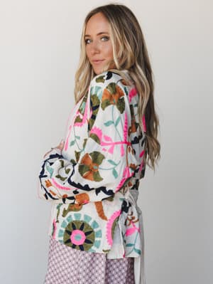 A model looks back over her shoulder in a richly embroidered white jacket layered over a lavender print skirt — the kind of piece that does all the talking so you don't have to.