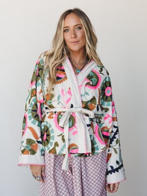 A richly embroidered wrap jacket in cream with bold floral and botanical motifs in hot pink, olive, teal, and rust — belted at the waist with wide sleeves and a relaxed, artisan feel.