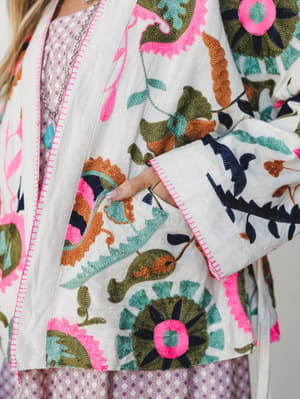 A close-up detail shot of a heavily embroidered cream jacket layered over a purple printed dress — the kind of piece that stops people mid-sentence to ask where you got it.